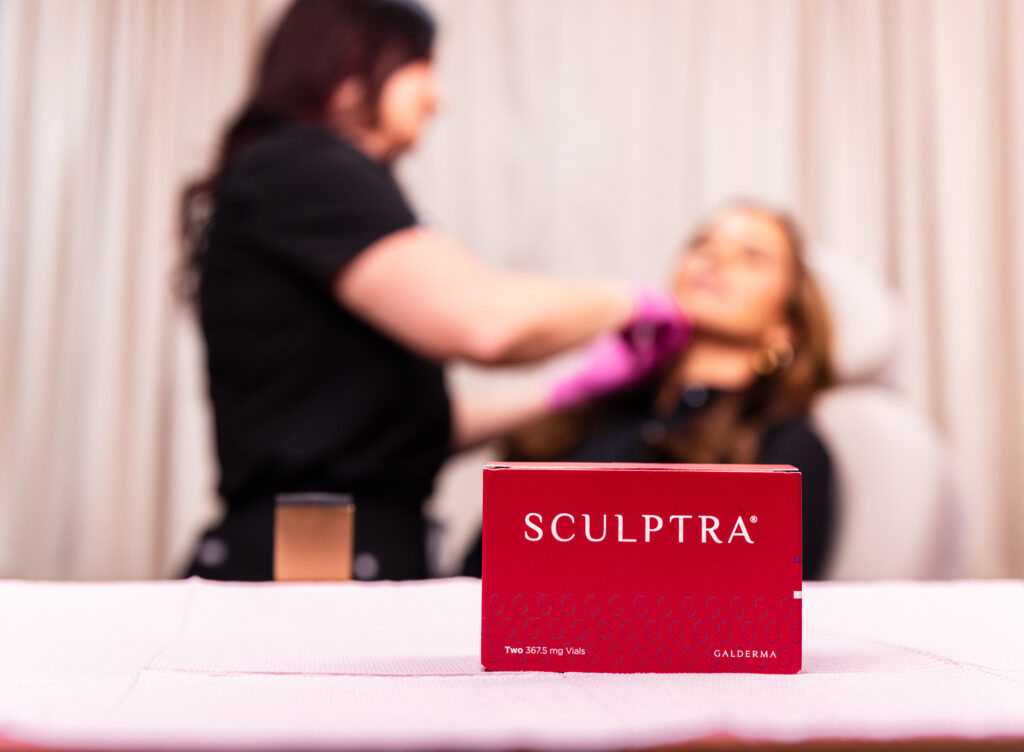 Front and center is a box that contains units of Sculptra near Cairo, GA. In the background, out of focus, a provider at Harmonized Aesthetics administers a Sculptra injection.