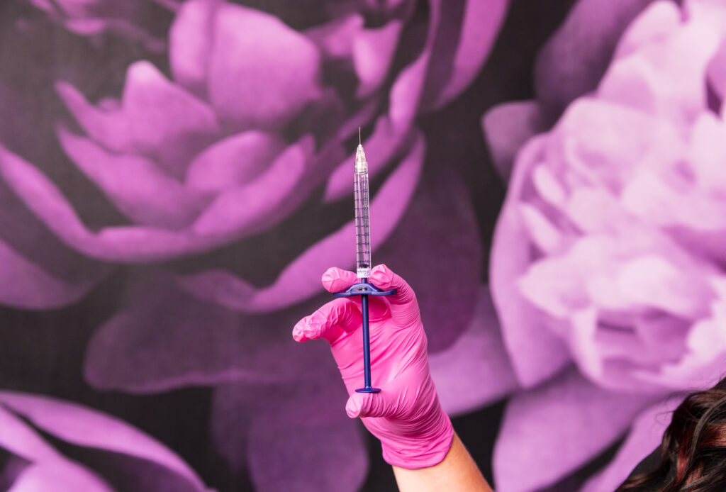 A surgical-gloved hand holds a syringe of dermal fillers, a wall with purple flower wallpaper in the background. Dermal fillers are used for tear trough filler in Thomasville, GA, to restore lost volume under the eyes.